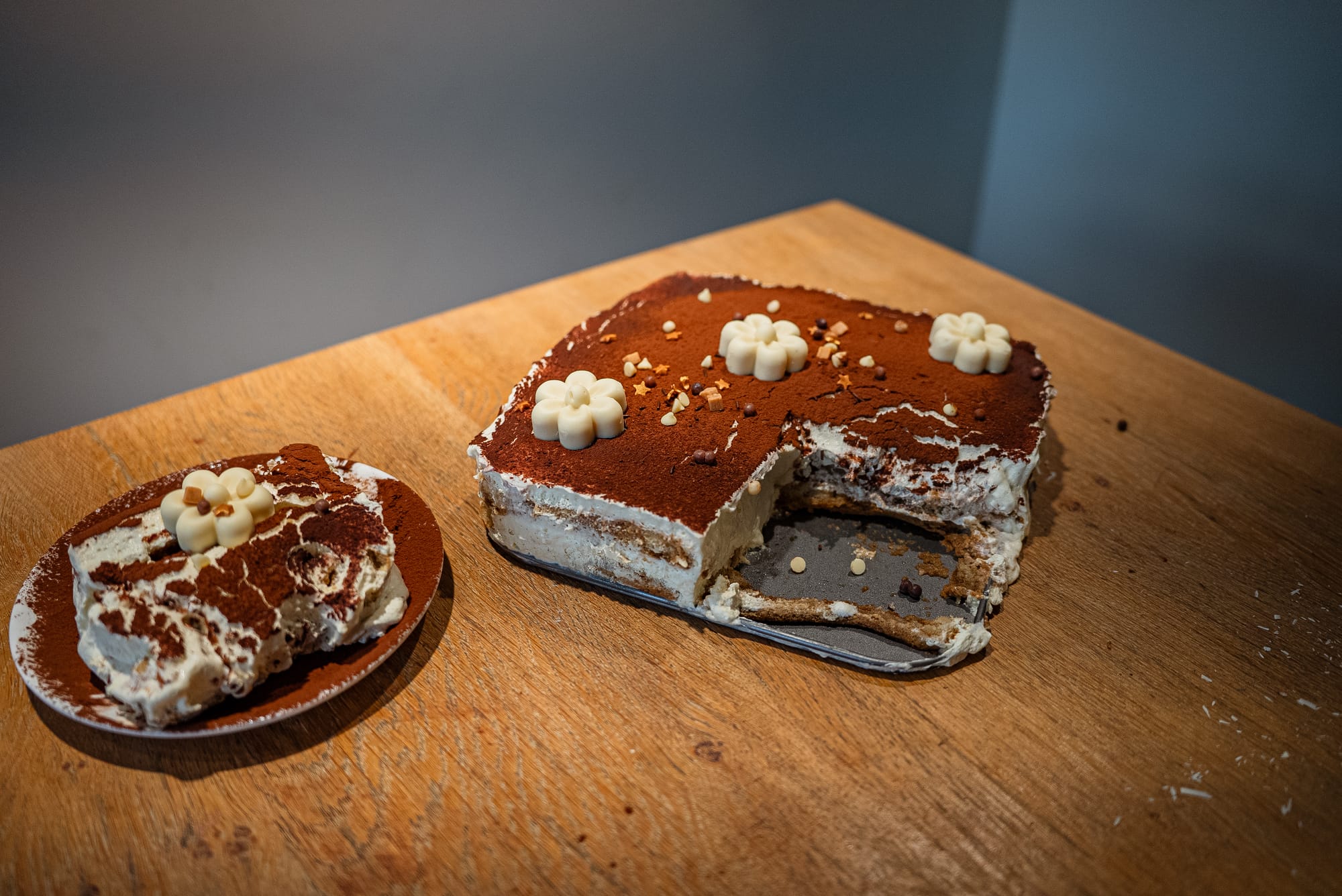Cut tiramisu slab decorated with white-chocolate flowers, served beside a plated slice
