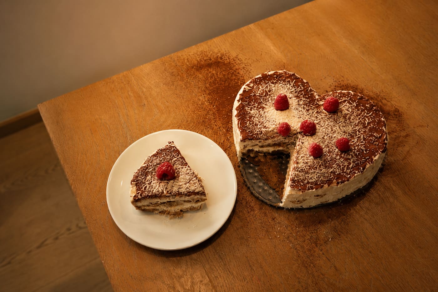 Heart-shaped tiramisu topped with raspberries beside a plated slice, photographed from above