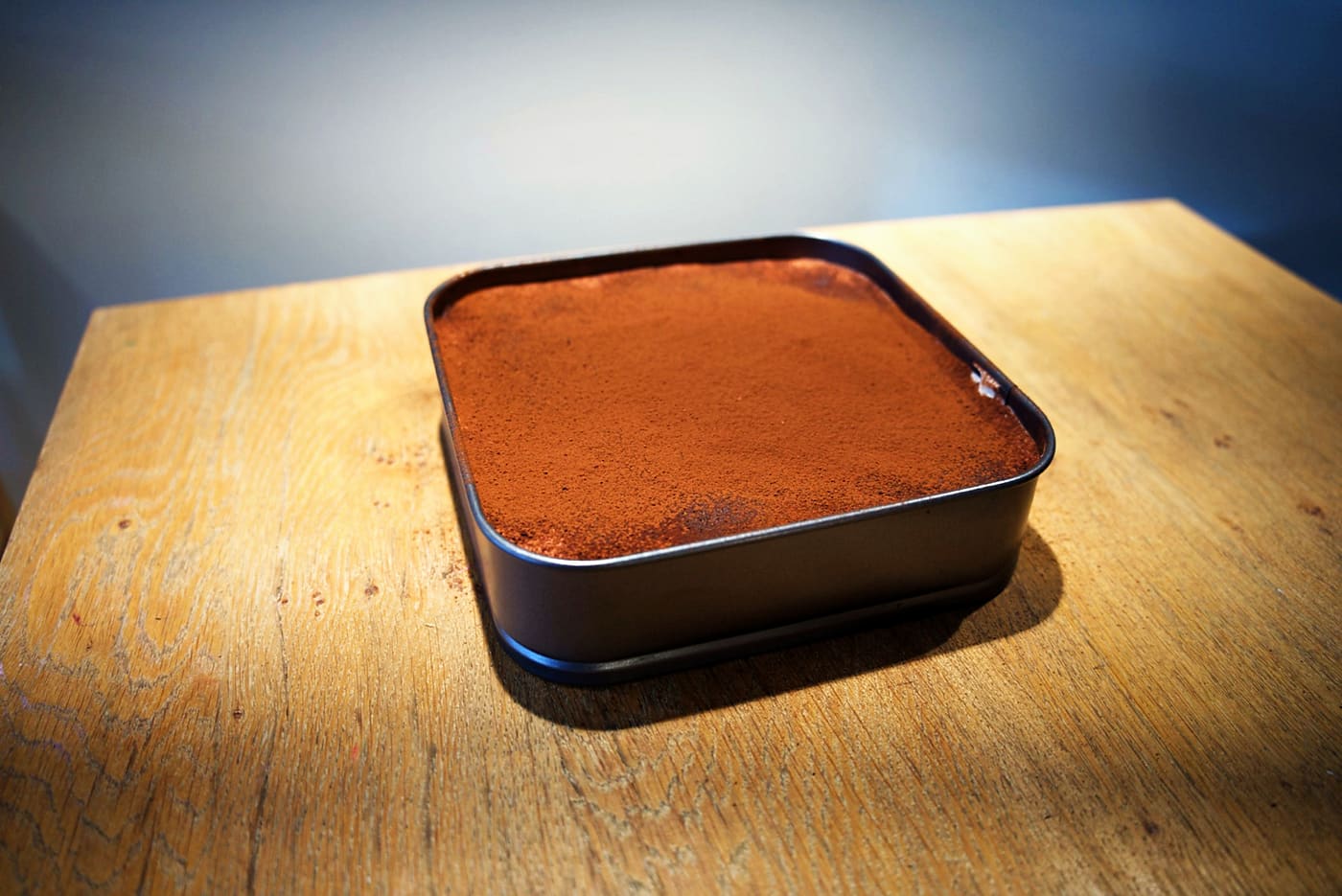 Cocoa-dusted tiramisu sharing slab in its tin on a wooden table