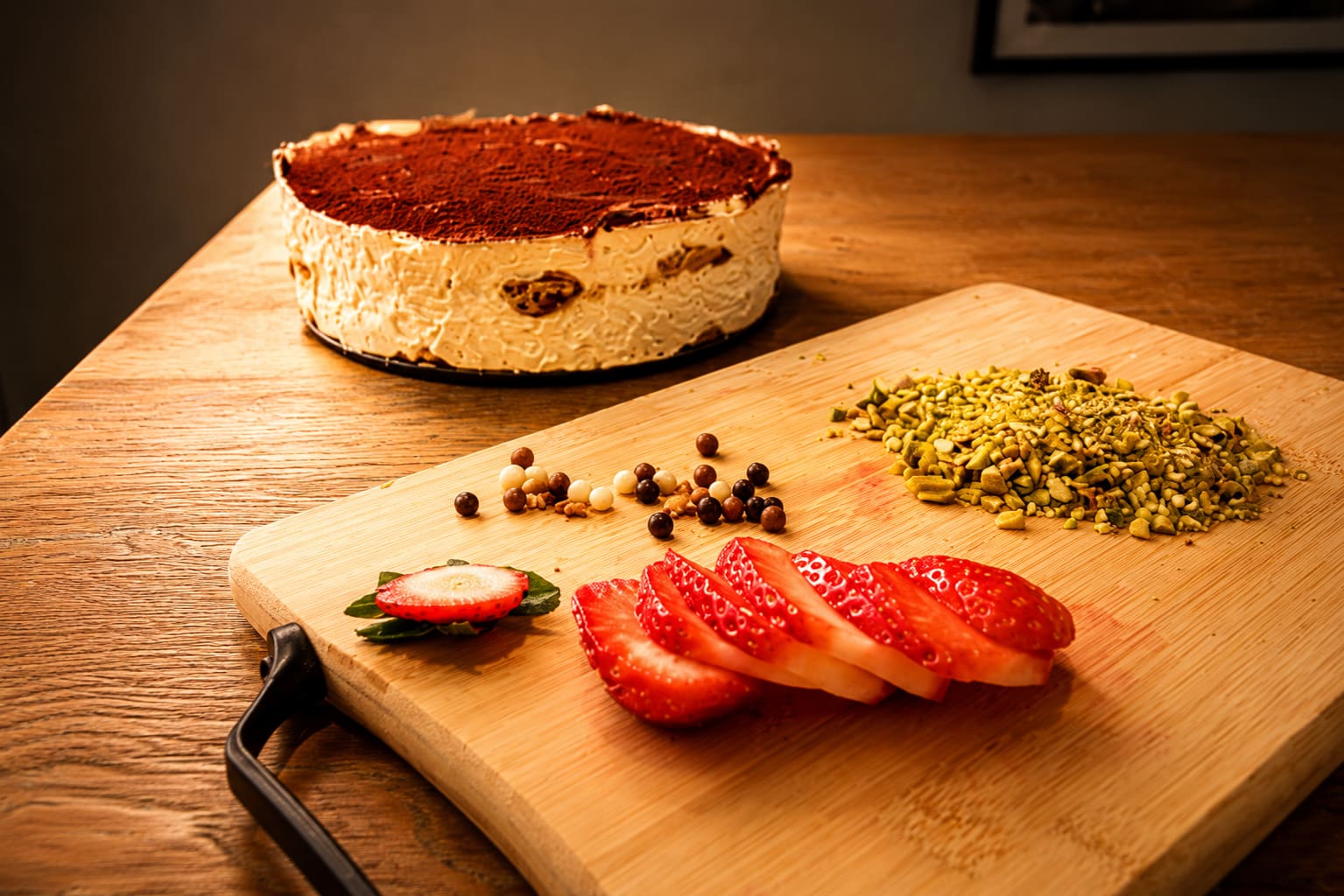 A Rai tiramisu round behind a chopping board of sliced strawberries, crushed pistachios and spices