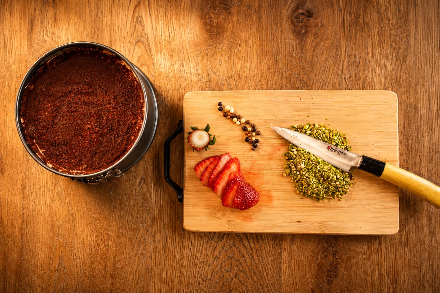 Overhead view of a tiramisu tin beside a chopping board of sliced strawberries and crushed pistachios